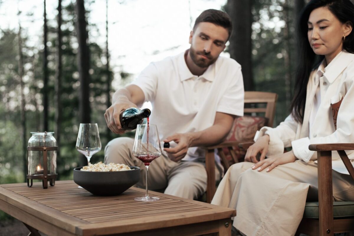 A couple relaxing in a forest setting, enjoying wine and popcorn at a wooden table.